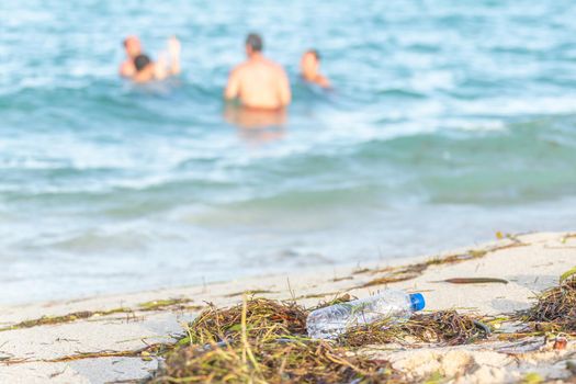 close up image of empty plastic water bottle on dirty beach filled with seaweed on dirty sand beach with people in sea on background