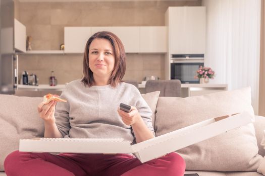 Beautiful woman eating pizza and watching TV holding remote control at home