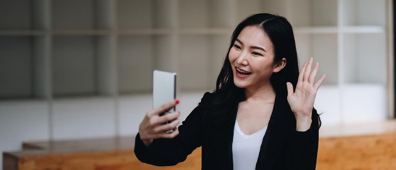 Happy young asian woman holding phone looking at screen waving hand video calling distance friend online in mobile chat app using smartphone videochat application.