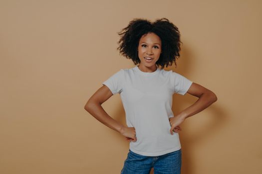 Studio portrait of beautiful confident african american female wearing casual stylish clothes keeping hands on hips and looking at camera with confidence, isolated on sand color background