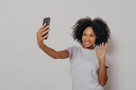 Happy young female waving at camera while having video call with best friend on modern smartphone, young cheerful african woman expressing positiveness while talking with followers in social media