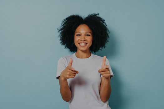 Positive friendly dark skinned lady making finger gun gesture and smiling at camera, happy young african female in white tshirt being happy to meet best friend, isolated over blue background
