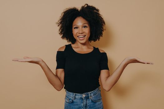 Unsure african female model in with curly hair shrugging shoulders with uncertainty with clueless and confused look while posing alone isolated over beige background, dressed in casual outfit