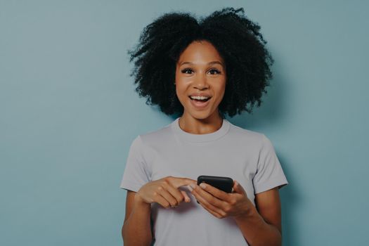 Happy dark skinned girl holding modern smartphone and chatting with friend online or using mobile application, pointing at device screen and looking at camera with excited face expression