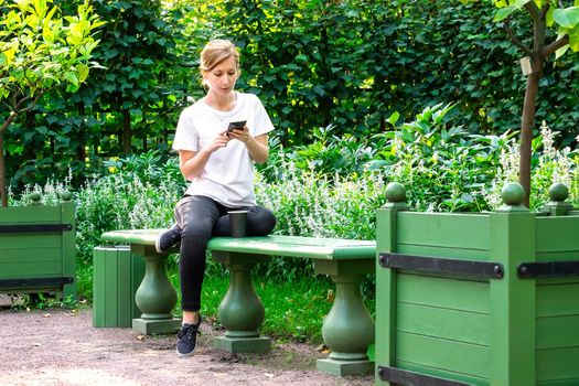 Young pretty blonde in white T-shirt sits on bench in beautiful garden with her legs curled up and holds smartphone in her hands. Woman looks at smartphone screen and reads message from social media.