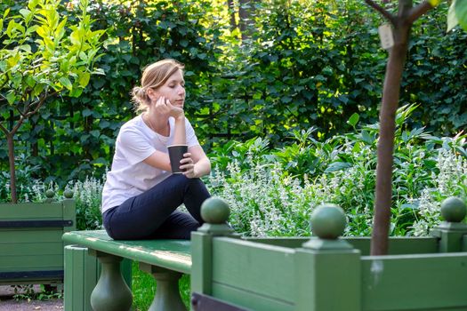 Young pretty blonde in white T-shirt sits on bench in park garden with her legs tucked in and her head propped on her hand, holding an eco-friendly paper cup with coffee in her hand. Selective focus