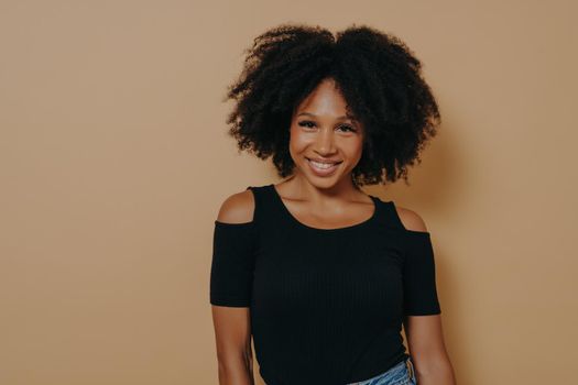 Smiling afro american woman with curly hair standing against beige background, has happy and positive face expression, young african female in casual outfit looking at camera with tender smile