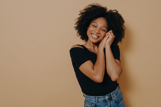 Happy cute 20s african woman with curly hair keeping hands near face and eyes closed, smiling pleasantly while dreaming of something good, isolated over beige background. Human emotions concept