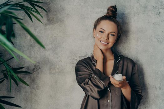 Image of cheerful woman in silk pajama smiling while applying face cream, posing with jar of moisturizing lotion in her hand, isolated over concrete wall background. Beauty and skin care concept