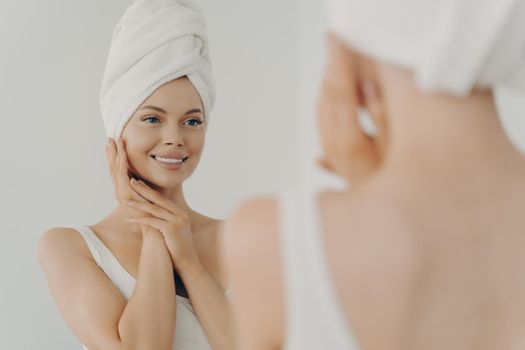 Beautiful happy woman after applying make up looking in mirror and smiling while standing in bathroom, pretty young female with white towel on head gently touching face. Beauty and skin care concept