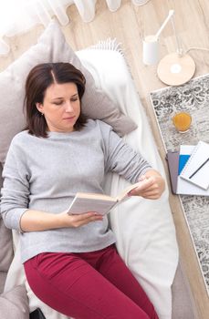 Young woman reading a book as she relaxes on her back on a comfortable sofa, view from above