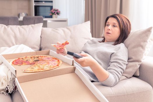 Beautiful woman eating pizza and watching TV holding remote control at home