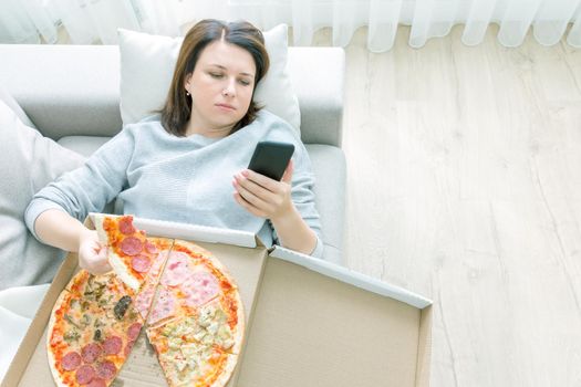 Sad woman eating pizza and holding phone laying on a couch at home