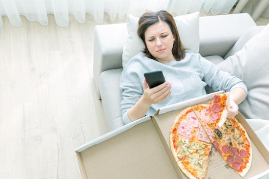 Sad woman eating pizza and holding phone laying on a couch at home