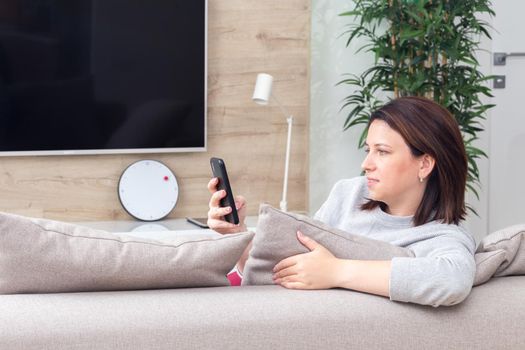 Young woman looking at her smart phone and smiling while sitting on the couch at home