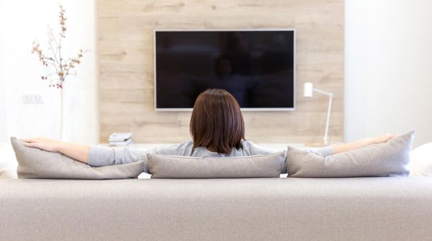 Young woman watching TV in living room, back view