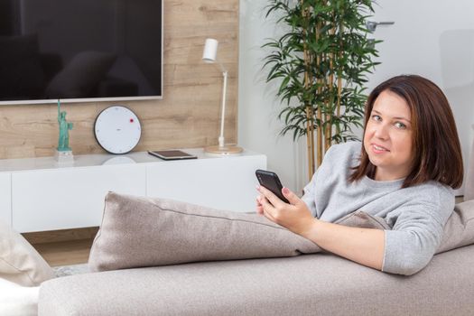 Young woman looking at her smart phone and smiling while sitting on the couch at home