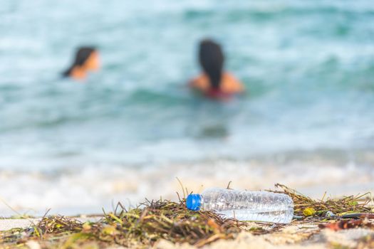 close up image of empty plastic water bottle on dirty beach filled with seaweed on dirty sand beach with people in sea on background