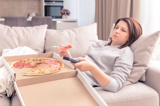 Sad woman eating pizza and holding phone laying on a couch at home