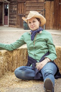 Cowgirl woman smiling happy sitting next to haystack wearing cowboy hat. Beautiful young Caucasian girl in countryside