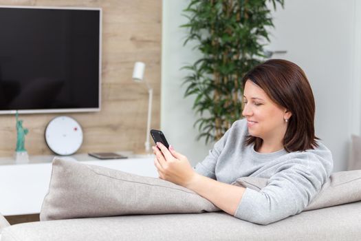 Young woman looking at her smart phone and smiling while sitting on the couch at home