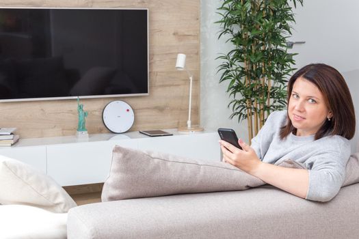 Young woman looking at her smart phone and smiling while sitting on the couch at home