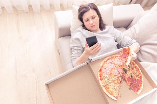 Sad woman eating pizza and holding phone laying on a couch at home