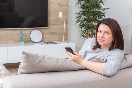 Young woman looking at her smart phone and smiling while sitting on the couch at home