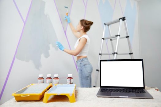 Young woman painting wall with paint roller and using masking tape. Laptop on table with white copy space mockup