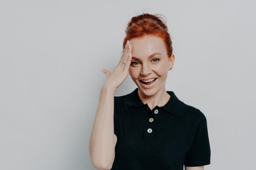 Portrait of pretty redhaired woman with hair in bun dressed in black t-shirt polo forgetting something, slapping forehead with palm and smiling at camera while posing against grey wall. Memory concept
