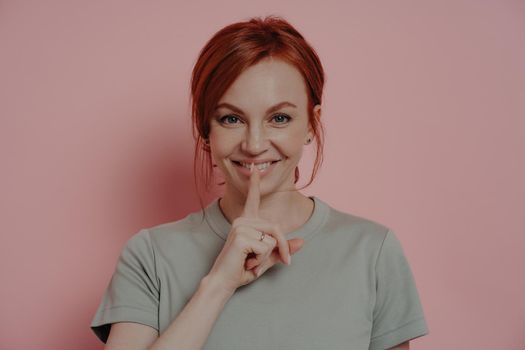 Young beautiful smiling red-haired woman telling secret information, posing isolated over pink wall, keeping forefinger over lips and smiling at camera, asking keep it in secret. Secrecy and privacy