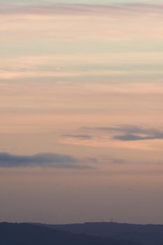 Vertical hazy sunset. Horizon under warm and faded pastel colours. Gently clouded sky above the forested horizon with protruding telecom towers.