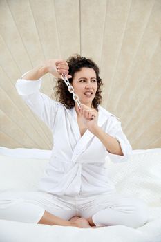 Young beautiful brunette curly woman sitting in lotus pose in a white bed holding metal chain