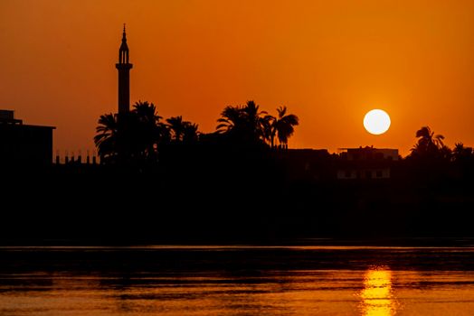 View across large wide river Nile in Egypt through rural countryside landscape with beautiful orange sunset and mosque minaret architecture