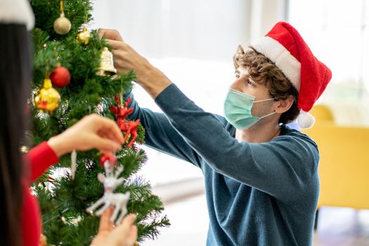 Close up Caucasian man with hygiene mask decorate the Christmas tree with his couple in his home for celebrate the festival during pandemic of Covid-19 and new normal lifestyle.