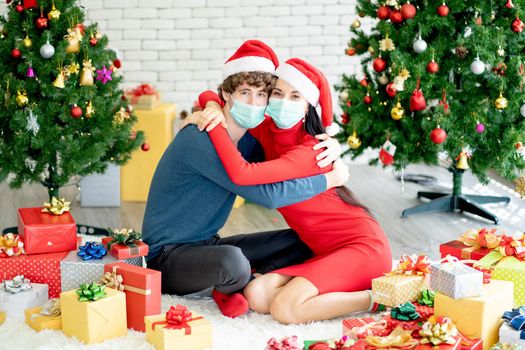 Couple man and woman with hygiene mask sit on floor and hug together with many Christmas decorations during pandemic of Covid-19.