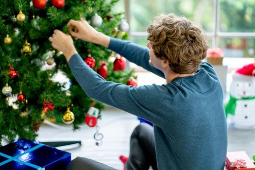 Back of man decorate the Christmas tree in the room of his house and he put many gift box or present around area the he stay.