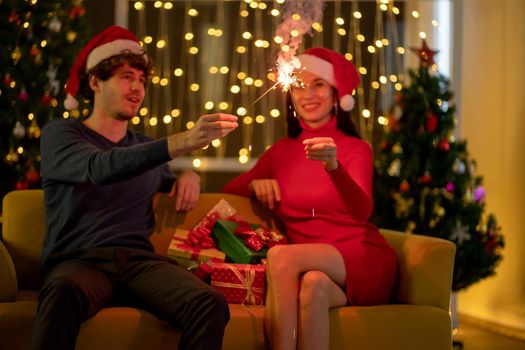 Couple man and woman with Christmas costume have fun with sparklers in living room of their house at night with fairy lights as background.