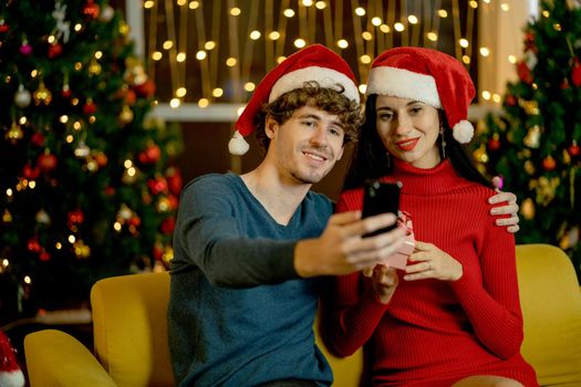 Couple man and woman use mobile phone to selfie video or picture of their good moment during Christmas festival at home with the decoration in background.