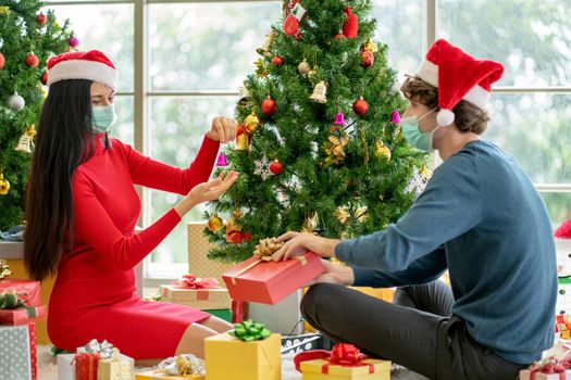 Couple man and woman with hygiene mask discuss and help to decorate Christmas tree together in room of their house.