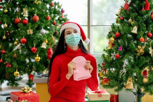 Woman with hygiene mask hold pink knit hat and look happy with sitting near Christmas tree and gift box for celebrating during pandemic of Covid-19.