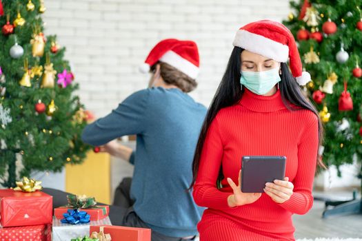Caucasian woman with hygiene mask choose the gift from online shop via tablet for celebration of Christmas during Covid-19 pandemic and new normal lifestyle to stay at home.