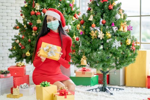 Portrait of Caucasian woman with hygiene mask sit on carpet with many giftbox and also hold one of giftbox with look at camera for Christmas during pandemic of Covid-19 with new normal lifestyle.