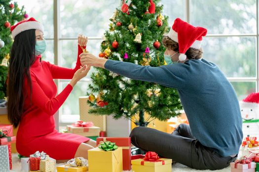 Couple man and woman with hygiene mask discuss and help to decorate Christmas tree together in room of their house.