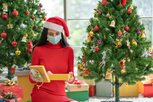 Caucasian woman with hygiene mask hold and look at giftbox, she also sit near Christmas tree.