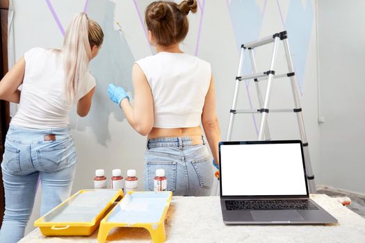 Young woman painting wall with paint roller and using masking tape. Laptop on table with white copy space mockup