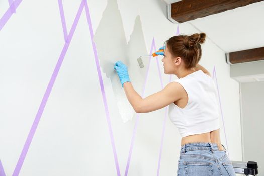 Young woman painting wall with paint roller while standing on a ladder. Masking tape is used to make painted objects