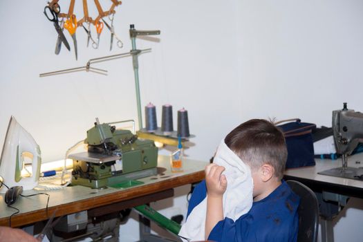 Boy is cleaning face with a white towel after a haircut at home in home sewing room. Part of living at home during lockdown. Stay at home.