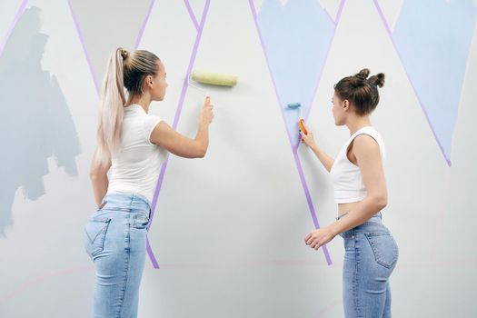 Young woman painting wall with paint roller while standing on a ladder. Masking tape is used to make painted objects