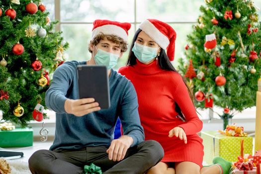 Couple man and woman with hygiene mask use tablet to celebrate christmas with other people also their family during pandemic of Covid-19 and new normal lifestyle.